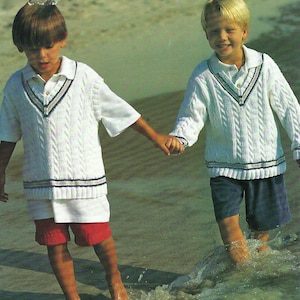 Può includere: Due giovani ragazzi che indossano maglioni di lana a treccia bianca con bordini blu navy camminano lungo una spiaggia sabbiosa. Il ragazzo a sinistra indossa pantaloncini rossi e il ragazzo a destra indossa pantaloncini blu.