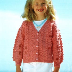 Puede incluir: Una niña pequeña lleva una chaqueta de punto de color rosa hecha a crochet con una camisa blanca de botones debajo. Está sonriendo y lleva un collar azul claro con un colgante en forma de corazón.