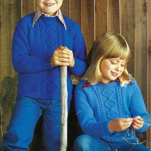 Puede incluir: Dos niños con suéteres azules de punto de cable. El niño sostiene un palo de madera y la niña sostiene un trozo de cuerda. Ambos están sentados frente a una pared de madera.