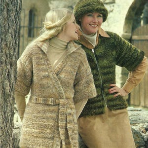 May include: Two women wearing knitted outerwear. One wears a tan wrap coat with a belt. The other wears a green zip-up jacket and matching hat. Both outfits are textured and appear to be made of a thick yarn, suitable for cooler weather.