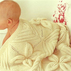 May include: A cream-colored, knitted baby blanket with a floral design. The blanket features large, three-dimensional flower appliqués and is draped over a baby. The blanket is made of soft yarn and appears cozy.