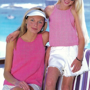 May include: Two people wearing pink knitted tank tops and white shorts. One person is wearing a white visor. The other person is wearing sunglasses. The background shows a blue ocean and sky.