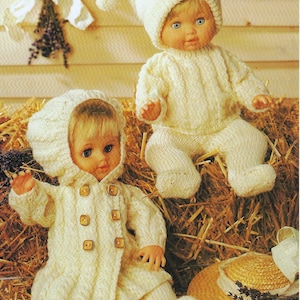 May include: Two knitted doll outfits in white, one with a hooded jacket and the other with a sweater and trousers. Both outfits feature a cable knit pattern and are displayed on a bed of straw.