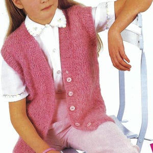 May include: A pink knitted waistcoat with a button fastening. The waistcoat has a V-neck and is sleeveless. The model is wearing a white shirt and pink trousers.