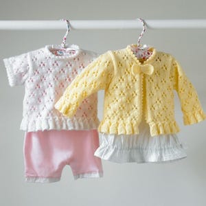 Op de afbeelding: Twee gehaakte baby-outfits, een wit met roze bies en een geel met witte bies. Beide outfits hebben een kantpatroon en hangen aan hangers.