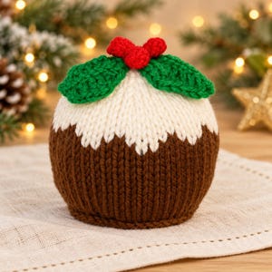 Op de afbeelding: Een gebreide kerstpudding-decoratie met een donkerbruine basis, een wit bovengedeelte dat op glazuur lijkt, en een decoratieve bovenkant bestaande uit twee groene gehaakte blaadjes en drie kleine rode besjes.