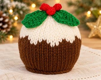 May include: A round, knitted Christmas pudding decoration featuring a dark brown base, a white top section resembling icing, and a decorative topper consisting of two green crocheted leaves and three small red berries.