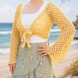 May include: A yellow crochet bolero with long sleeves tied at the front. The bolero is worn over a white tank top and paired with a green floral wrap skirt. The background shows a beach with the ocean and palm trees.