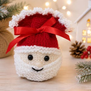 May include: A small, round, knitted Santa Claus pouch featuring a red hat with a white ruffled trim, a white beard, and a face with two black bead eyes and a stitched black mouth. A bright red satin ribbon is tied in a bow around the top section.