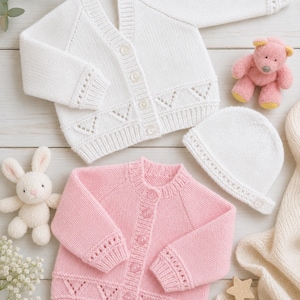 May include: A white knitted cardigan with buttons and eyelet details, a pink knitted cardigan with buttons and eyelet details, a white knitted hat, a pink plush bear, a white plush rabbit, and two wooden star shapes on a light wooden surface.