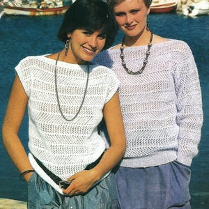 May include: Two women wearing white crocheted tops with short sleeves. The woman on the left is wearing a blue denim skirt and a silver necklace. The woman on the right is wearing a light blue skirt and a beaded necklace.