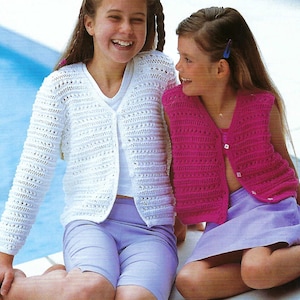 Può includere: Due ragazze che indossano abiti all'uncinetto fatti a mano. Una ragazza indossa un cardigan bianco a maniche lunghe con chiusura a bottoni, abbinato a pantaloncini lavanda. L'altra ragazza indossa un gilet rosa all'uncinetto e una gonna lavanda. Entrambe sorridono.