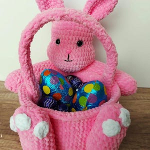 Op de afbeelding: Roze gehaakte konijnenmand met witte voetjes en een handvat. De mand is gevuld met in folie verpakte chocolade paaseieren.