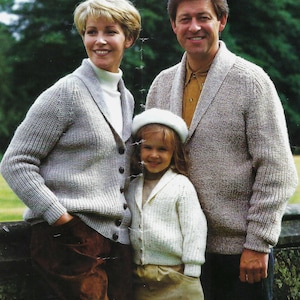 May include: A family of three wearing knitted cardigans. The adults are wearing beige cardigans with a shawl collar. The child is wearing a white cardigan with a shawl collar. The child is wearing a white beret.