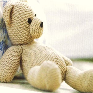 Puede incluir: Un oso de peluche de punto beige con ojos negros. El oso está tumbado sobre una superficie blanca.