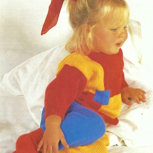 Op de afbeelding: Een kind draagt een kleurrijke gebreide outfit. De outfit bestaat uit een rode top met gele accenten, een blauwe legging en rode sokken. Een rode strik siert het blonde haar van het kind. De outfit heeft een zak aan de voorkant.