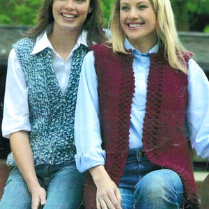 May include: Two women wearing knitted vests. The woman on the left is wearing a blue and white vest, while the woman on the right is wearing a burgundy vest. Both women are wearing white shirts and blue jeans.