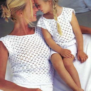 Puede incluir: Una madre y su hija llevan tops de crochet blanco a juego con un delicado patrón de calado. Los tops tienen un escote redondo y son sin mangas.