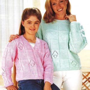 May include: Two people wearing knitted sweaters. One is wearing a light pink cardigan with diamond cutouts, paired with blue jeans. The other is wearing a light blue sweater with a similar diamond pattern, paired with white pants.