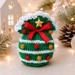 May include: A green crocheted drawstring pouch featuring white horizontal stripes, a red ribbon bow, a gold star embellishment, and small red and gold pom-poms. The pouch has a scalloped white crocheted top edge and a rounded base shape.