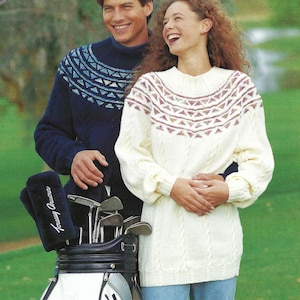 May include: A couple stands on a golf course, both wearing patterned sweaters. The man wears a navy blue sweater with a blue and white design. The woman wears an off-white sweater with a cable knit design and a geometric pattern. A golf bag is in the foreground.