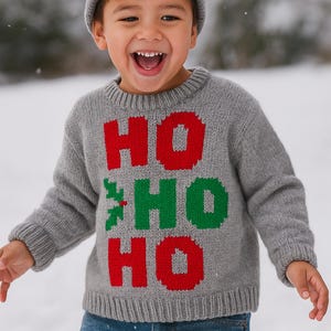 May include: A child in a grey knitted jumper with the words "HO HO HO" in red and green. The child is also wearing a grey beanie and blue jeans. The background is a snowy scene.