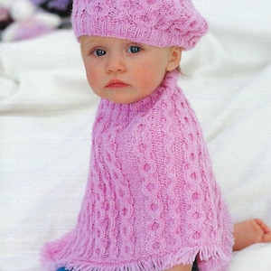 May include: A baby in a pink knitted beret and poncho. The poncho has a fringed hem and is worn with blue jeans. The knitted set features a textured pattern, creating a cosy and stylish look for the child.