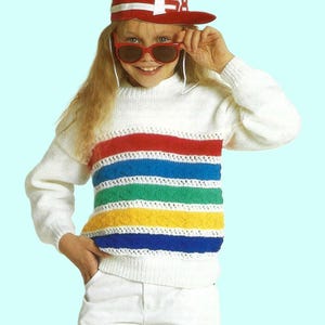 May include: A white sweater with rainbow stripes and a white crocheted trim. The sweater has a crew neck and long sleeves. The model is wearing a red baseball cap with the number 58 on it.
