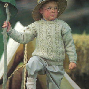 Può includere: Un bambino piccolo che indossa un maglione di lana beige chiaro a trecce, pantaloni kaki e un cappello di paglia con un nastro blu. Sta tenendo un bastone di legno e guardando a destra.