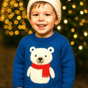 May include: A young child wearing a blue sweater with a white polar bear design and a red scarf. The child is also wearing a white knit cap. The background features blurred Christmas tree lights.