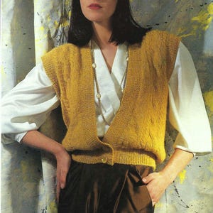 May include: A woman in a mustard yellow knitted vest, white blouse, and dark brown skirt. The vest features a cable knit pattern and is buttoned at the waist. A gold necklace is worn. The background is a painted wall with grey, yellow, and white colours.