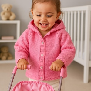 May include: A young child wearing a pink hooded cardigan, smiling and pushing a pink doll's pram. The pram has a pink fabric seat and a doll inside. The background includes a cot and a shelf with a teddy bear.