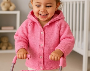 Puede incluir: Una niña sonriente con una rebeca rosa con capucha y botones, empujando un cochecito de muñecas rosa. El cochecito tiene un asiento de tela rosa y una muñeca dentro. El fondo incluye una cuna y una estantería con un osito de peluche.