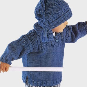 Puede incluir: Un suéter azul de punto con un gorro azul de punto a juego. El suéter tiene un patrón de punto de cable y un cierre de botón en el cuello. El gorro tiene un pompón en la parte superior.