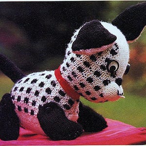 May include: A black and white knitted dalmatian dog toy with a red collar. The dog has a large, round, black eye and a black nose.
