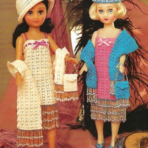 May include: Two dolls wearing crocheted dresses and matching shawls. The doll on the left is wearing a white dress and shawl with a white hat. The doll on the right is wearing a pink dress and a blue shawl. Both dolls are holding crocheted purses.