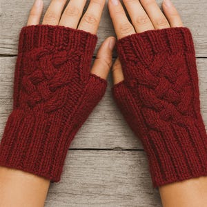 May include: A pair of burgundy fingerless gloves with a cable knit design. The gloves are made of a soft, knitted material and feature a ribbed cuff. The cable knit pattern is centered on the back of the hand.