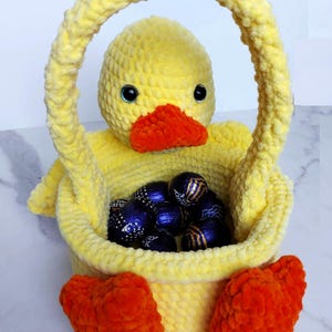 May include: A yellow crocheted Easter basket shaped like a duck with orange feet and a handle. The basket is filled with blue and gold wrapped chocolate eggs.