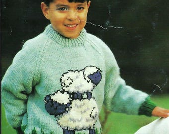 May include: A young boy wearing a light green knitted sweater with a white sheep design. The sweater has a crew neck and long sleeves. The boy is standing in a grassy field and is holding a white dog.