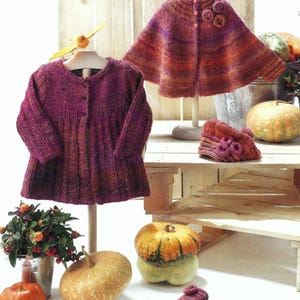 May include: A set of knitted baby clothes in shades of purple and orange. The set includes a cardigan, a cape, a hat, and booties. The items are displayed on a wooden pallet with pumpkins.