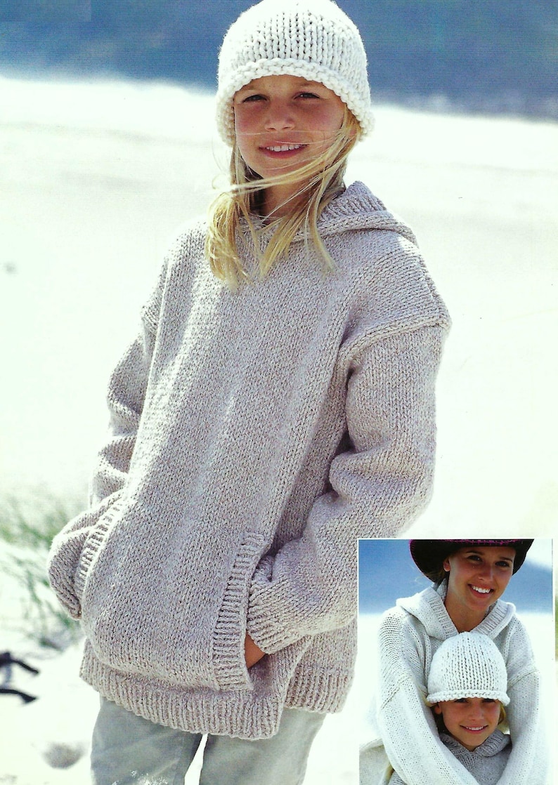 May include: A light gray hooded sweater with a pocket on the front. The sweater is made of a soft, knitted fabric. The model is wearing a white knitted hat.