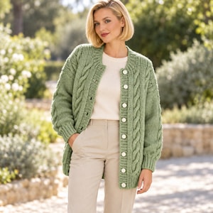 Puede incluir: Un cárdigan de punto trenzado en color verde salvia con cuello redondo, mangas largas con puños acanalados y cierre frontal de botones blancos. La prenda se lleva sobre una camiseta blanca lisa y unos pantalones de color beige claro.