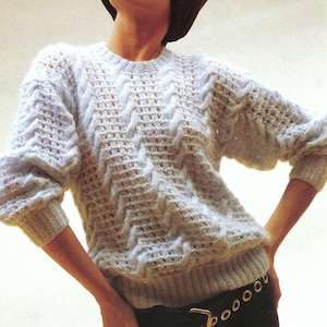 May include: A light blue knit sweater with a textured chevron pattern. The sweater has long sleeves and a ribbed hem and cuffs. The model is wearing a black belt with a gold buckle.