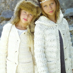 Könnte beinhalten: Zwei Mädchen tragen weiße Strickjacken mit Knöpfen. Ein Mädchen trägt eine braune Pelzmütze. Beide Jacken haben Taschen.