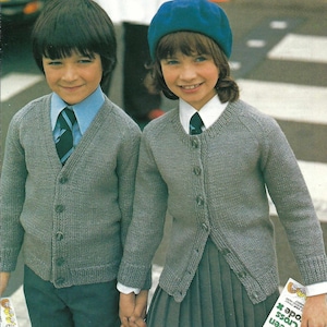 Puede incluir: Dos niños con cárdigans de punto gris con cierre de botones. El niño lleva una corbata a rayas azul y blanca. La niña lleva una boina azul y una camisa blanca con cuello y corbata azul. La niña sostiene un libro con el texto "The Green Cross Code" en la portada.