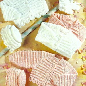May include: Two sets of knitted baby clothes, one in pink and one in white. Each set includes a cardigan, hat, and booties. The cardigans have a button closure and a scalloped edge. The hats are simple and the booties have a scalloped edge.