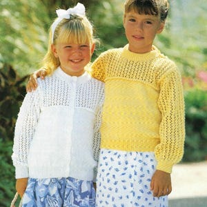 May include: Two children wearing knitted sweaters. The girl on the left is wearing a white sweater with a button closure and a blue and white patterned skirt. The girl on the right is wearing a yellow sweater with a white and blue patterned skirt.