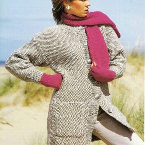 May include: A long, knitted cardigan with a button-up front and two pockets. The cardigan is a light grey colour and is worn with a pink scarf and matching fingerless gloves.