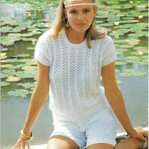 以下が含まれることがあります： 繊細な透かし模様の白いクロシェ編みセーター。セーターは半袖で丸首です。モデルはセーターに白いショートパンツとヘッドバンドを着用しています。