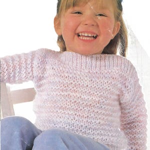 May include: A young girl wearing a pink knitted jumper with a light blue shirt underneath. She is smiling and sitting on a white chair.